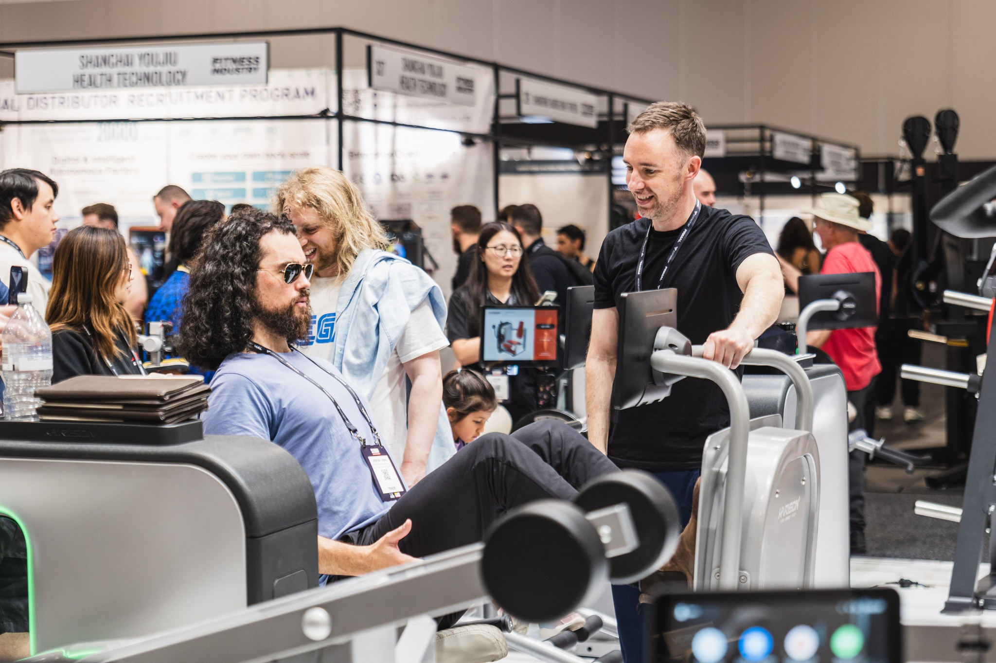 Exhibitor chatting with visitors trying a leg-press machine at AusFitness Expo, ICC Sydney
